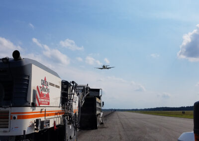 Asphalt milling machine on an airport runway with a plane taking off in the background.