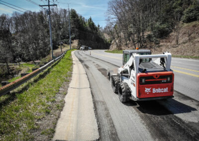 Road construction with a compact loader on a rural highway, surrounded by greenery and utility poles.