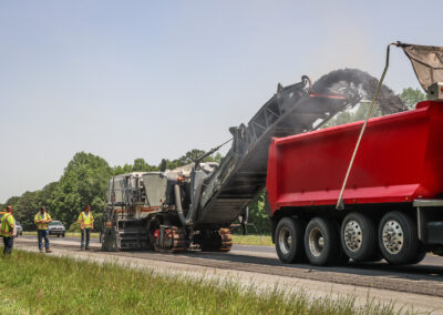 Workers overseeing an asphalt milling machine loading debris into a truck on a rural highway.