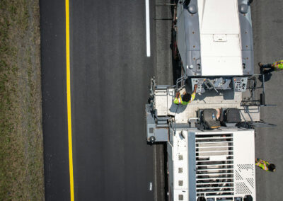 Top-down view of workers operating an asphalt milling machine on a highway next to a newly paved lane.