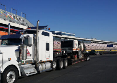 Heavy truck carrying an asphalt milling machine on a speedway track, surrounded by stadium bleachers.