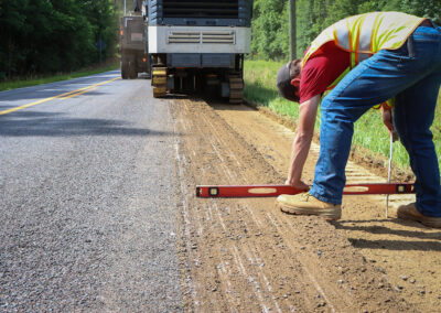 Construction worker measuring road surface with a level, asphalt milling machine in the background.