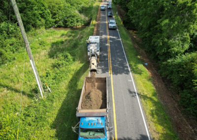 Aerial view of an asphalt milling machine and truck on a rural road, with cars lined up in the opposite lane.